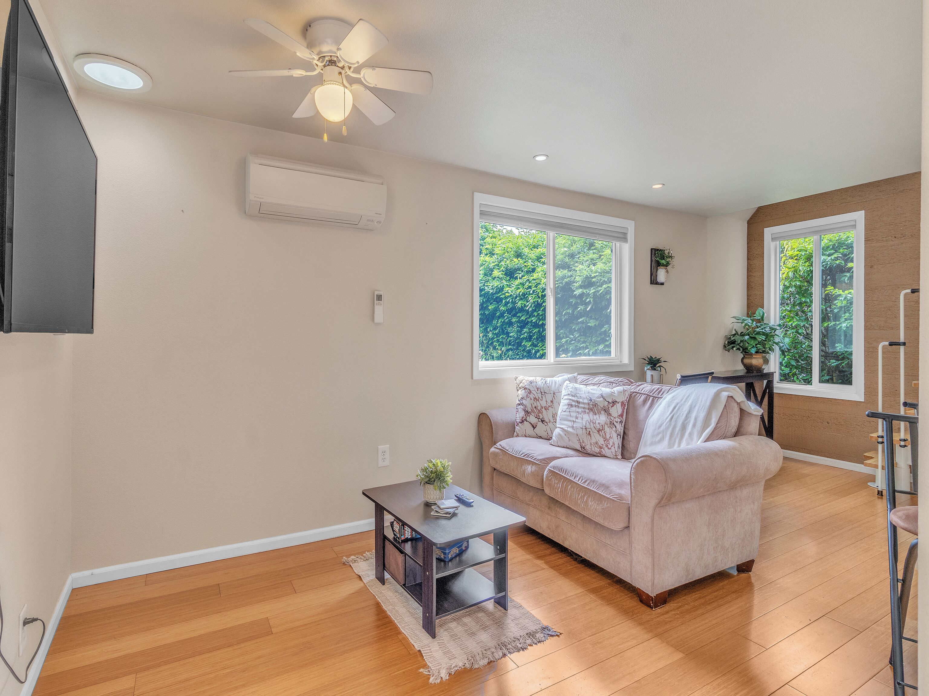 A living room with a beige couch, a coffee table, and a ceiling fan. There are two windows with a view of greenery outside.