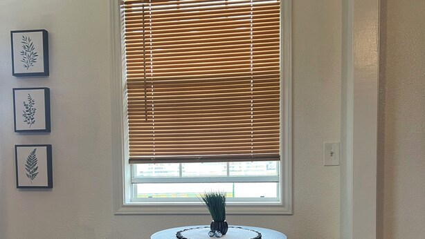 A small dining area with a round table and two stools, a window with blinds, and three framed botanical prints on the wall.