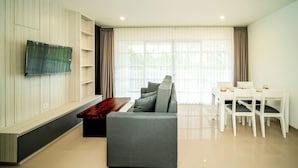 Ein modernes Wohnzimmer mit grauem Sofa, einem flachbildfernseher an der Wand, einem Esstisch mit Stühlen und einem großen Fenster mit durchsichtigen Vorhängen.