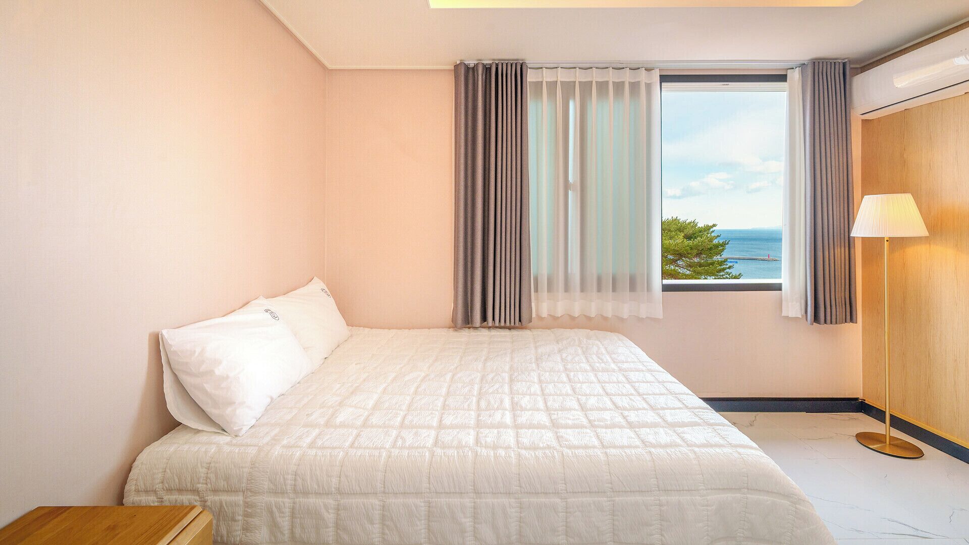 A hotel room with a bed, a window with curtains, a lamp, and a view of the sea.