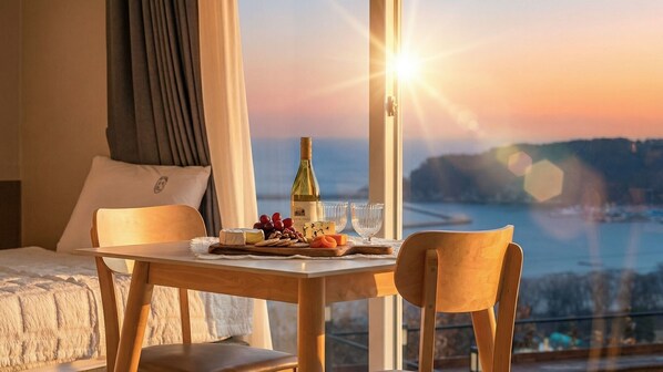 A hotel room with a dining table set for two, a bottle of wine, cheese, grapes, and a bowl of fruit.