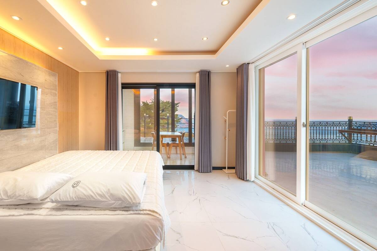 Una habitación de hotel moderna con una cama grande, un televisor, un balcón con vistas al mar y una puerta corrediza de vidrio que da a un espacio al aire libre.