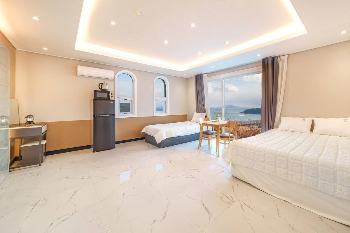 Habitación de hotel con cama, escritorio, refrigerador y ventana con vista al mar.