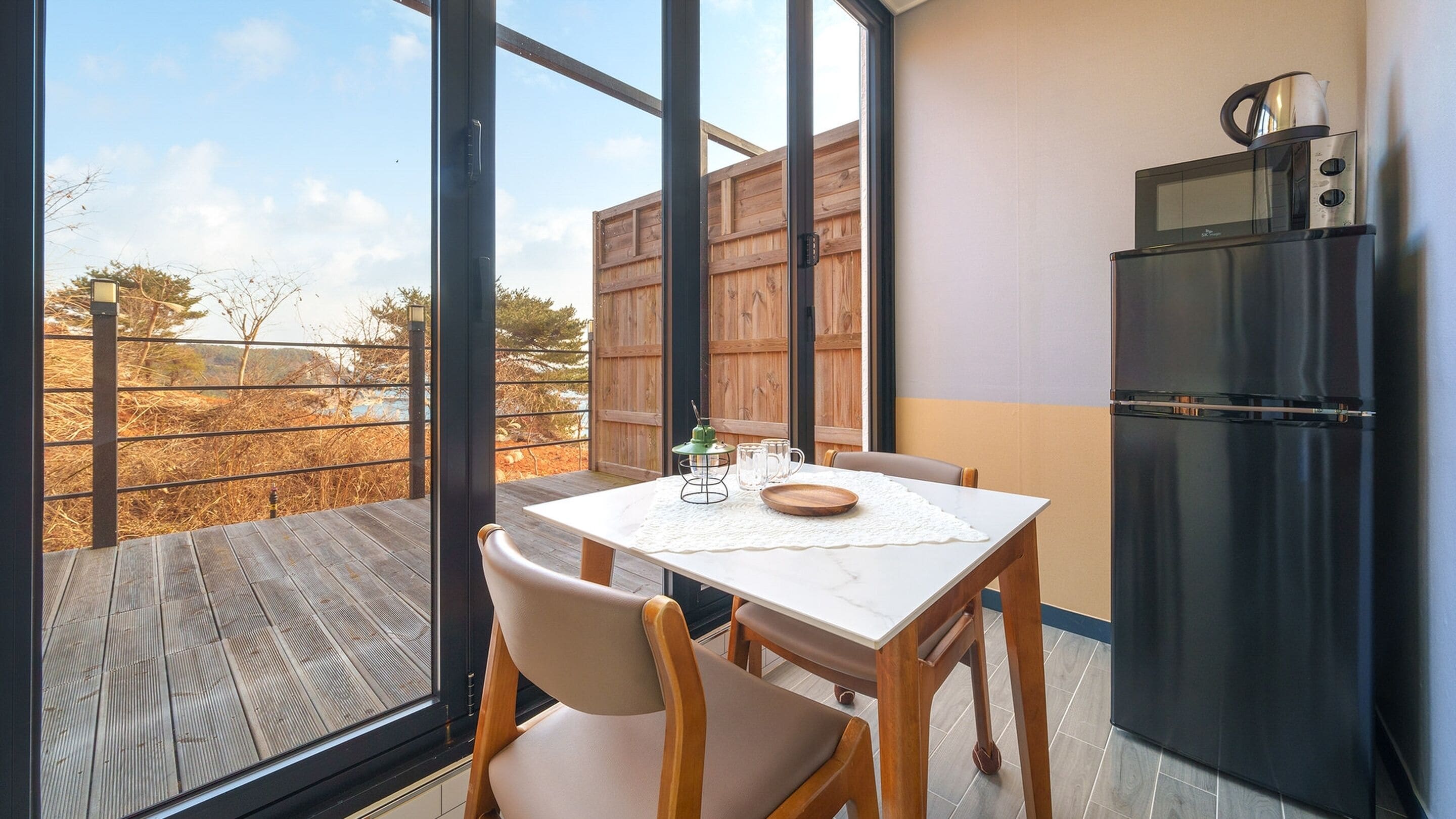 Een balkon met een tafel en stoelen, een koelkast, en uitzicht op bomen en een hek.