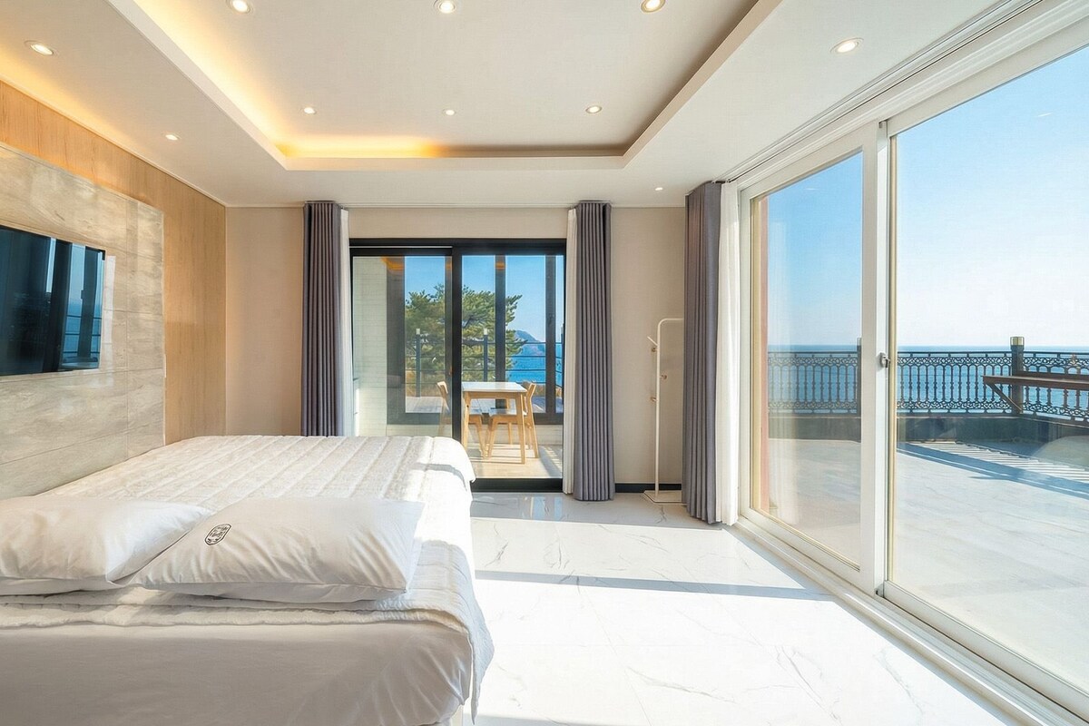 A modern hotel room with a large bed, a TV, and a balcony with a view of the sea.