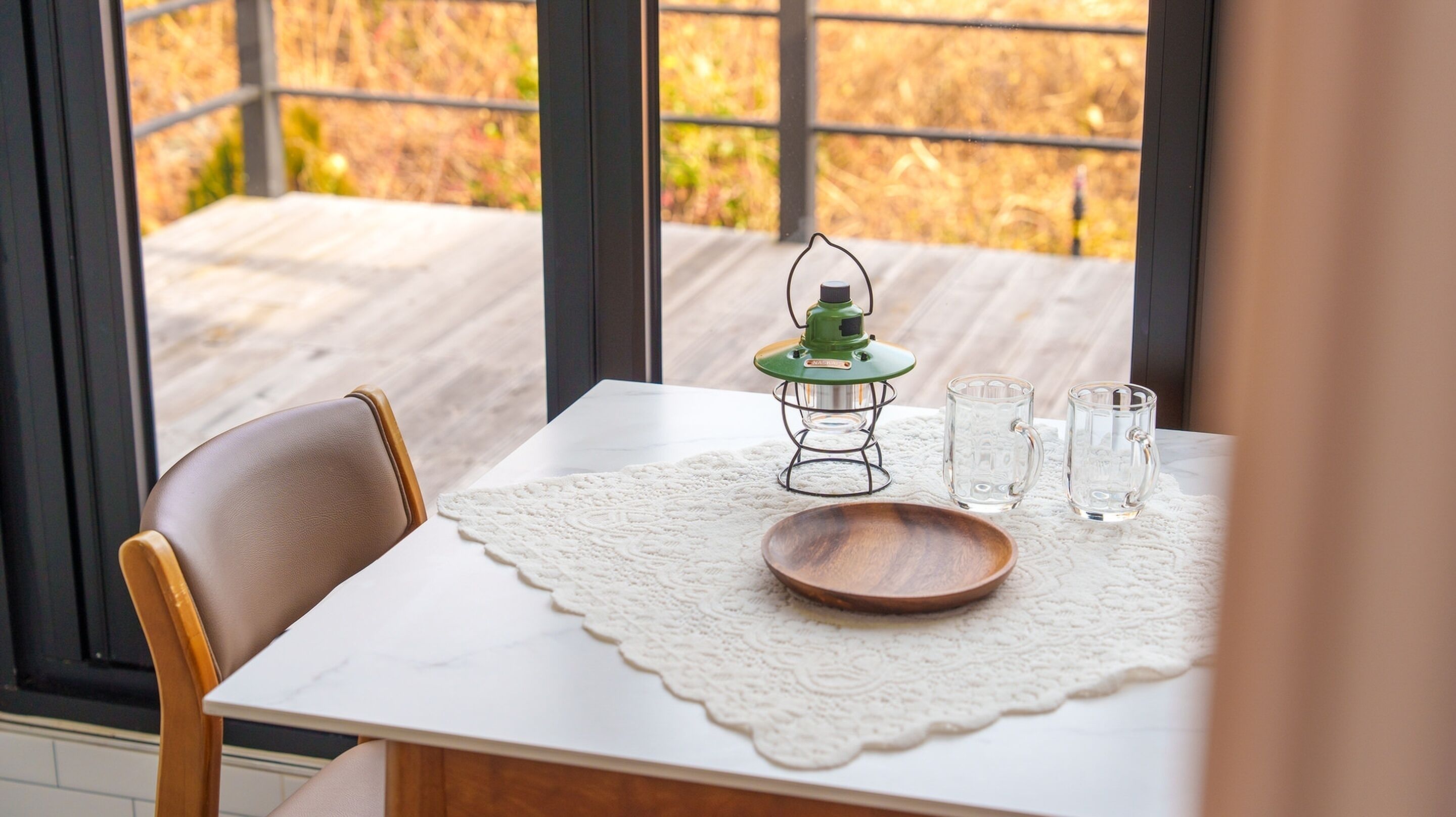 A table with a white lace tablecloth, a wooden plate, two clear glass mugs, and a green lantern.