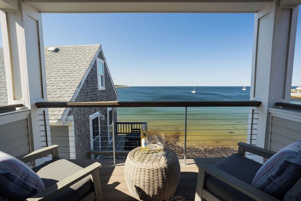 A balcony with a view of the sea, a wicker table with a bottle and glasses, and two chairs.