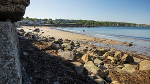 Une plage rocheuse parsemée de gros rochers, un ciel bleu dégagé et des maisons en arrière-plan.
