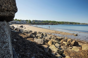 A rocky beach with large boulders, a clear blue sky, and houses in the background.