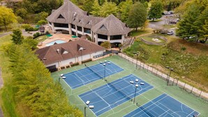 Vista aérea de un complejo de canchas de tenis con varias canchas, un edificio de diseño arquitectónico único y vegetación circundante.