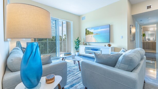 A modern living room with a large TV, grey sofas, a blue vase, and a view of the beach through the sliding doors.
