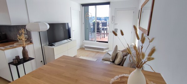 A modern living room with a flat-screen TV, wooden dining table, and a view of the outdoors through a sliding glass door.
