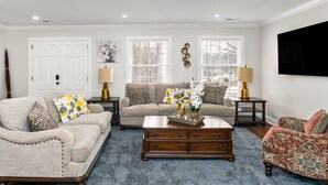 A living room with a beige sofa, a patterned armchair, a wooden coffee table, and a blue rug. There are two lamps on side tables, a vase with flowers, and a wall-mounted TV.