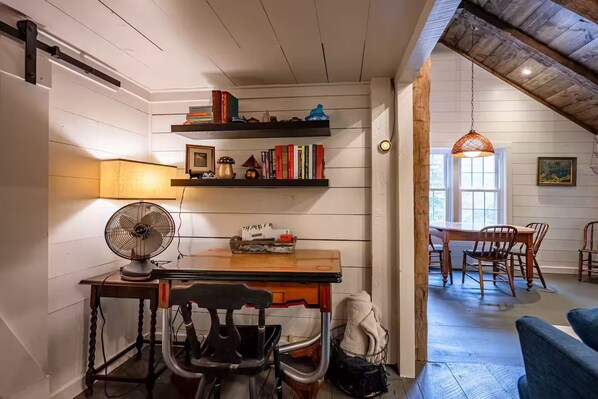 Una habitación acogedora con una mesa de madera, sillas, un ventilador y estantes con libros y objetos decorativos.