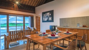 A dining area with a wooden table set for four, a kitchen area with granite countertops, and large windows offering a view of a green landscape. - The Manik Kalisada (Seririt)