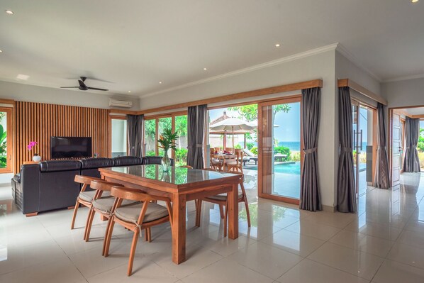 Uma sala de estar ampla com cozinha moderna, mesa de jantar e uma grande porta de vidro deslizante que dá para a piscina e vista para o mar.