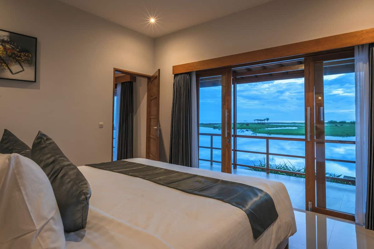 A bedroom with a large bed, a view of a water body, and a wooden ceiling.