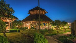 A thatched-roof pavilion with a covered outdoor seating area, surrounded by well-maintained gardens and a pathway leading to a building with a modern facade. - The Manik Kalisada (Seririt)