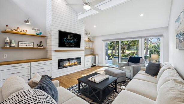 A modern living room with a fireplace, white walls, wooden shelves, and a large window offering a view of the outdoors.