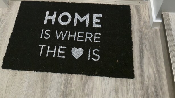 Eine Treppe mit Holzoptik und einem schwarzen Teppich am Fußende, auf dem der Schriftzug "HOME IS WHERE THE ♥ IS" steht.