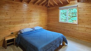 Habitación tipo cabaña de madera con cama, mesita de noche y ventana con vistas a árboles.