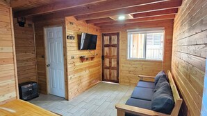 Interior de una cabaña de madera con un sofá, un televisor montado en la pared y una puerta que da a otra habitación.