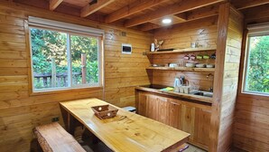 Interior de una cabaña de madera con una mesa de comedor, banco, zona de cocina y amplios ventanales con vistas al exterior.