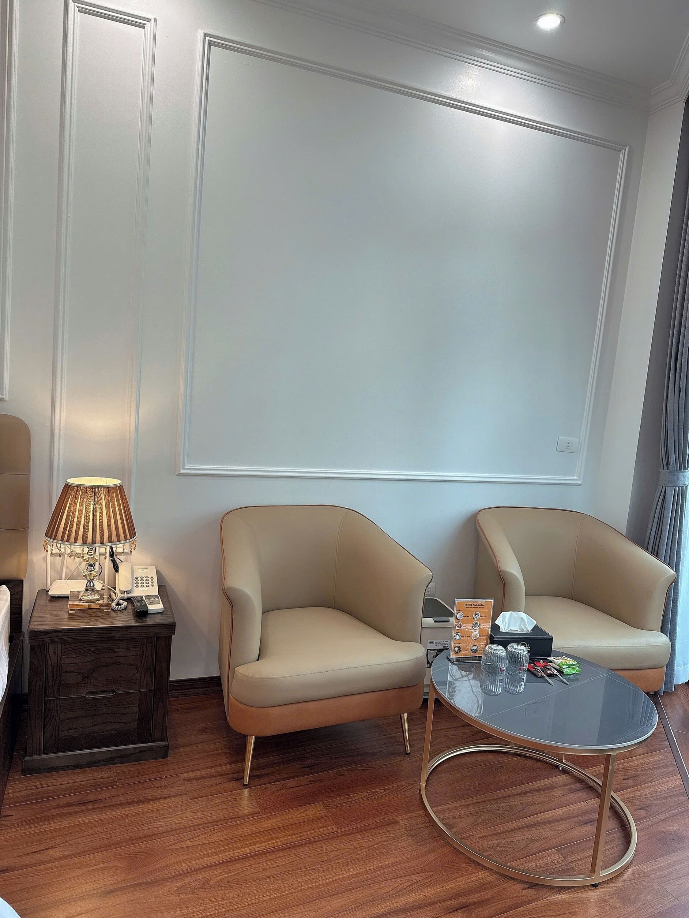 Ett hotellrum med två beige fåtöljer, ett litet träbord med en lampa, ett glasbord och en stor skärm som är monterad på väggen.