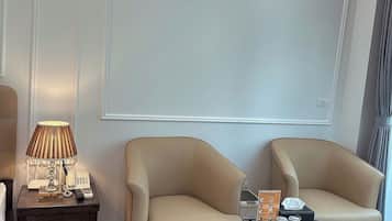Ett hotellrum med två beige fåtöljer, ett litet träbord med en lampa, ett glasbord och en stor skärm som är monterad på väggen.