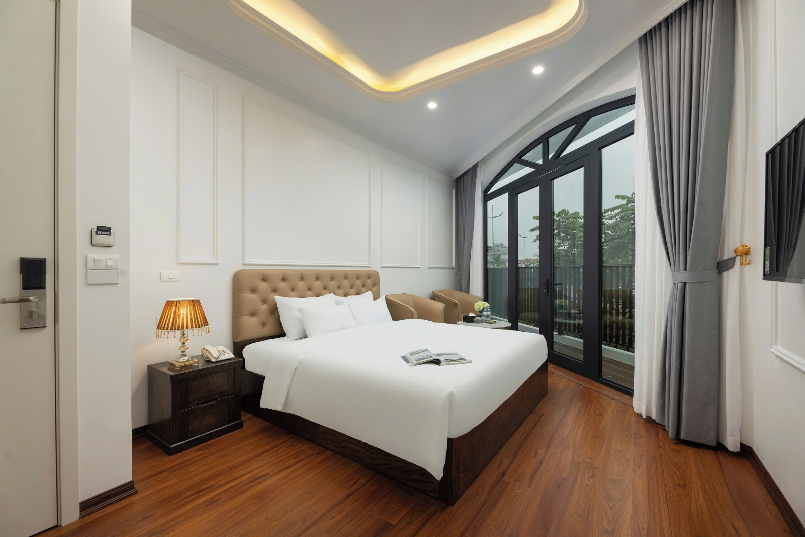 A modern hotel room with a large bed, a bedside table with a lamp, a nightstand, and a view of the outdoors through a large window.