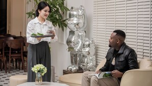 Una mujer sirve comida a un hombre en un interior moderno con una escultura plateada decorativa y una planta.