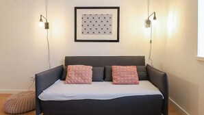 A modern bedroom with a dark sofa bed, two patterned pillows, a framed artwork above, and two wall-mounted lamps.