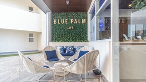 Un espacio al aire libre con sillas de mimbre y una mesa redonda, una pared con un fondo de follaje verde y un letrero que dice "BLUE PALM CAFE".
