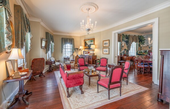 A spacious living room with a chandelier, red upholstered chairs, a wooden coffee table, and a large painting on the wall.