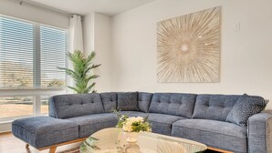 Uma sala de estar moderna com um sofá seccional cinza, uma mesa de centro de vidro e um grande quadro abstrato na parede.