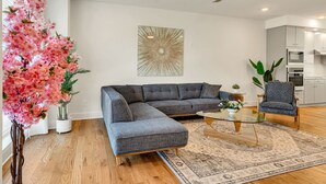 Uma sala de estar moderna com um sofá seccional, uma mesa de centro de vidro, um tapete estampado e uma árvore artificial rosa decorativa.
