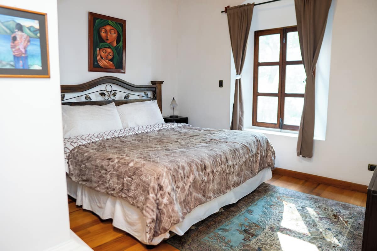A neatly made bed with a patterned bedspread, a bedside table with a lamp, a window with curtains, and a painting on the wall.