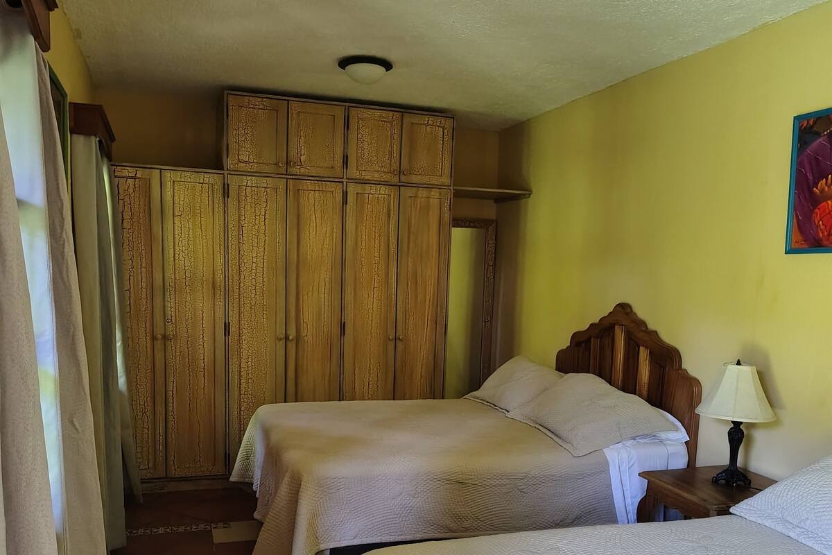 A room with two beds, a wooden wardrobe, a nightstand with a lamp, and a curtain.