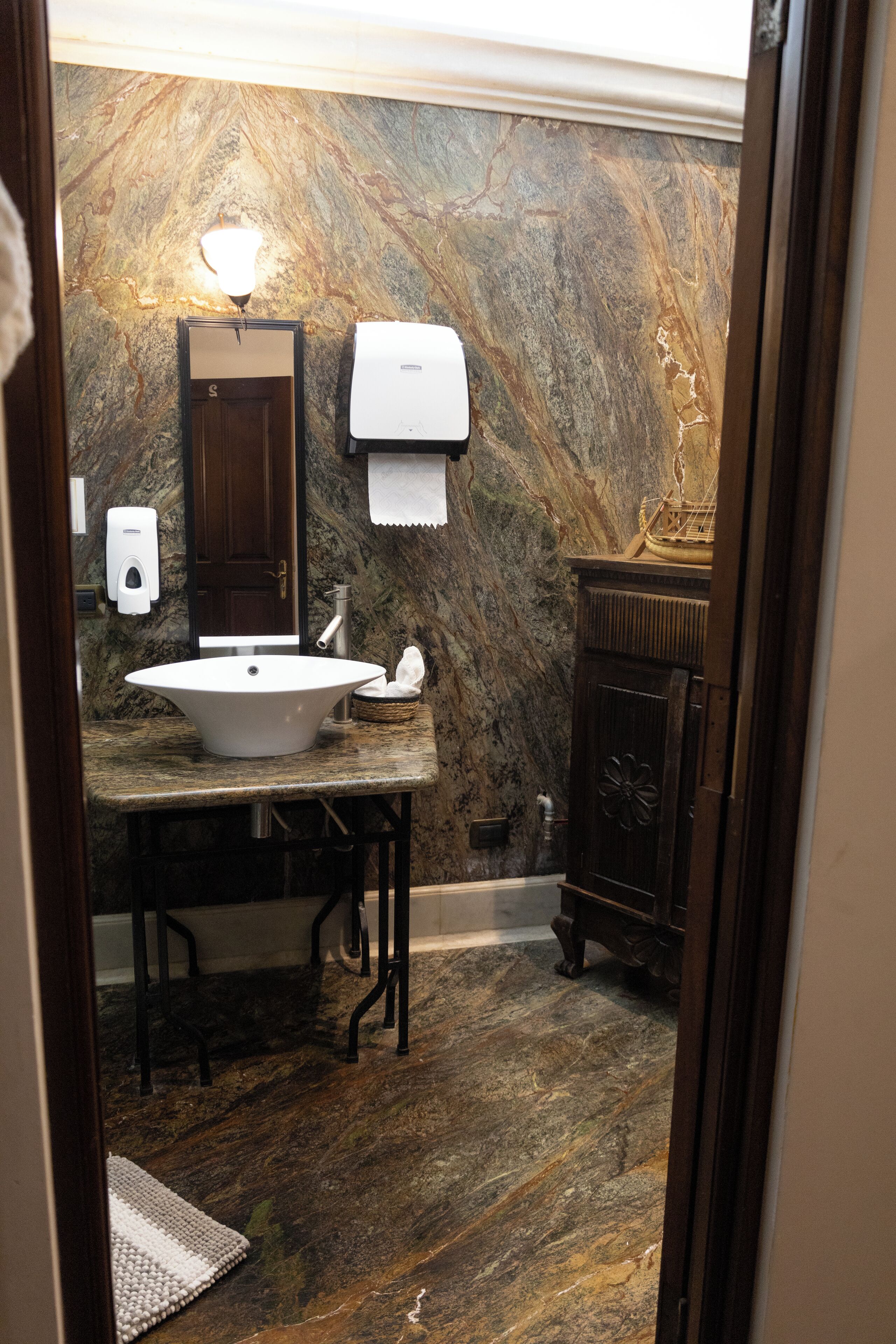 A bathroom with a marble wall, a sink, a mirror, a towel dispenser, and a wooden cabinet.