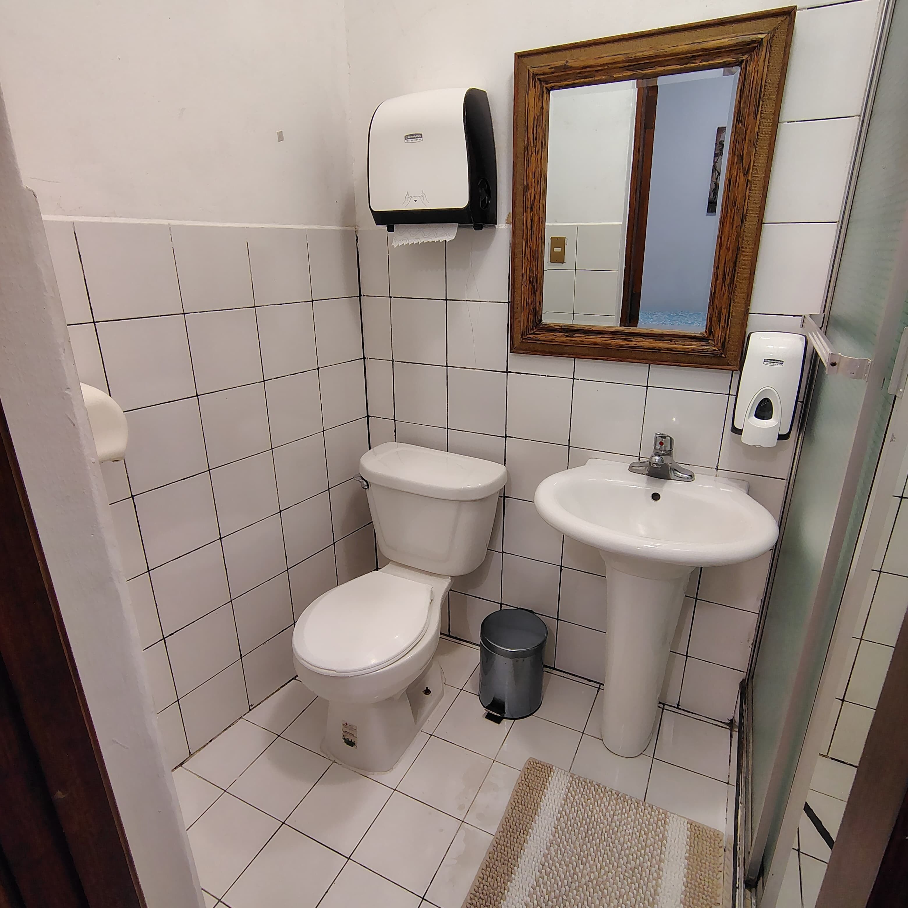 A small, clean bathroom with white tiles, a toilet, a sink, a hand dryer, and a trash can.
