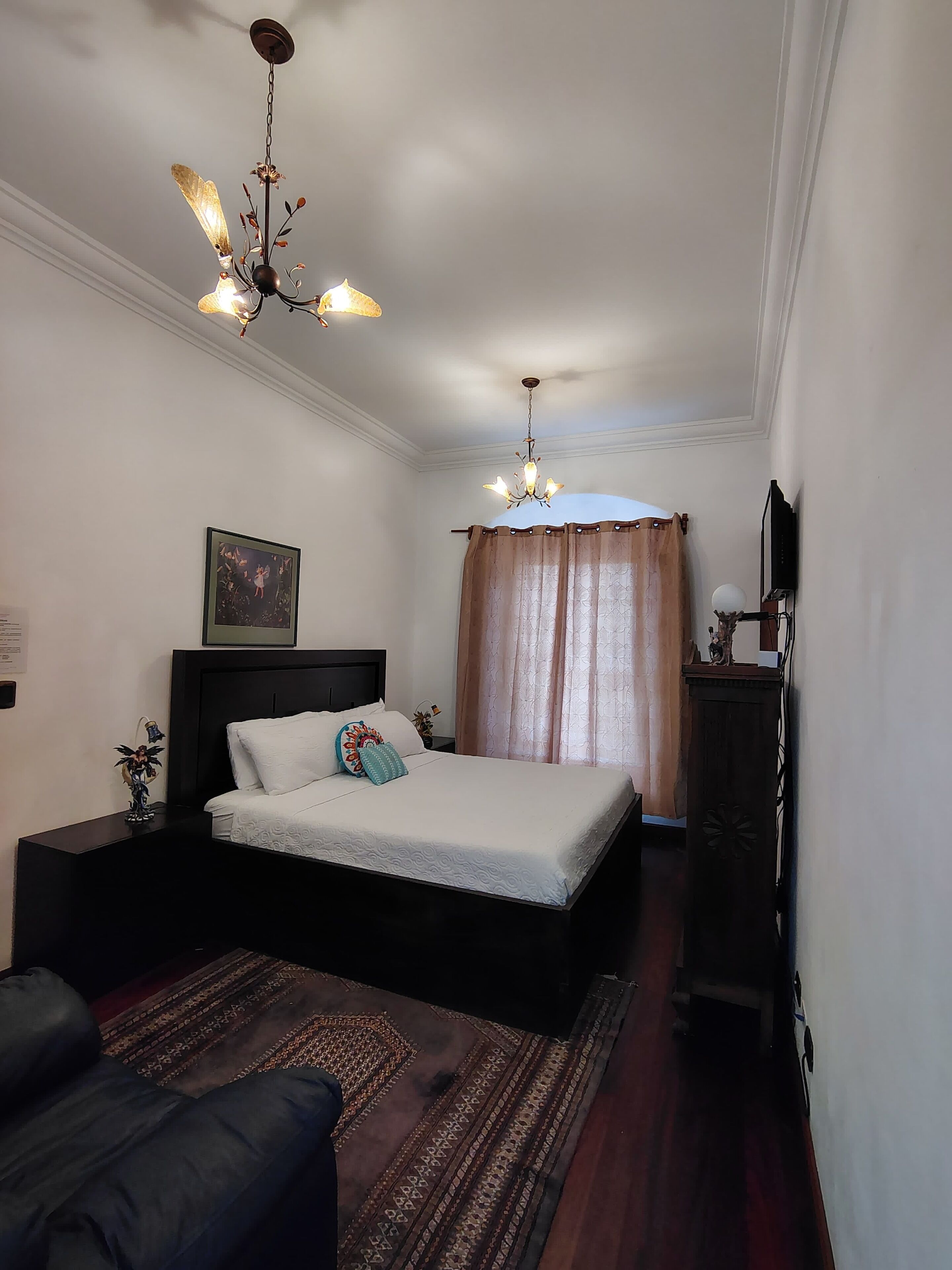 A bedroom with a bed, a nightstand, a dresser, a chandelier, a painting, and a curtain.