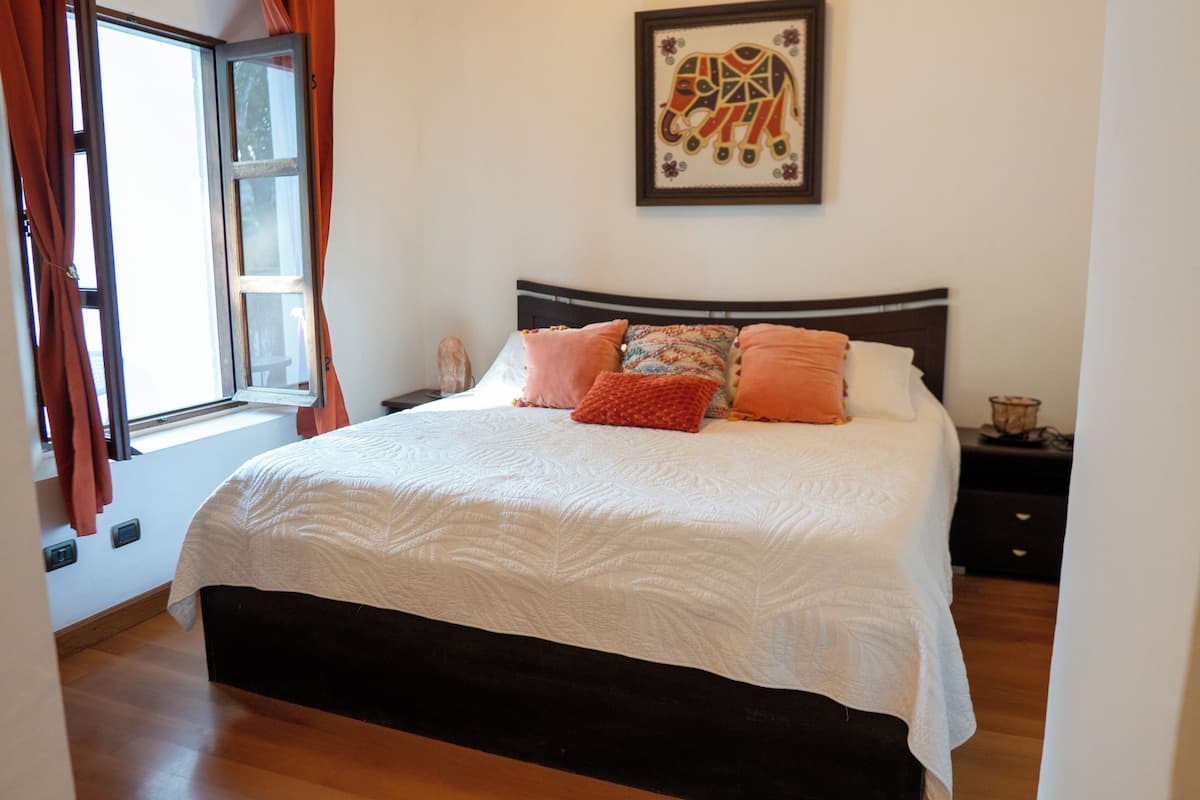 A neatly made bed with a white quilt and orange pillows, a framed elephant artwork on the wall, and a window with curtains.