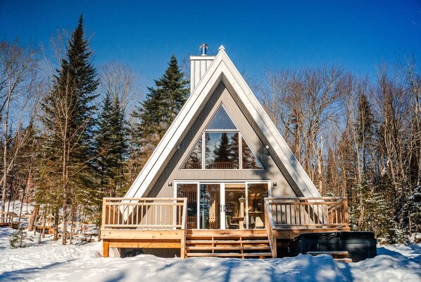 A triangular cabin with a wooden deck and large windows, surrounded by a snowy forest.