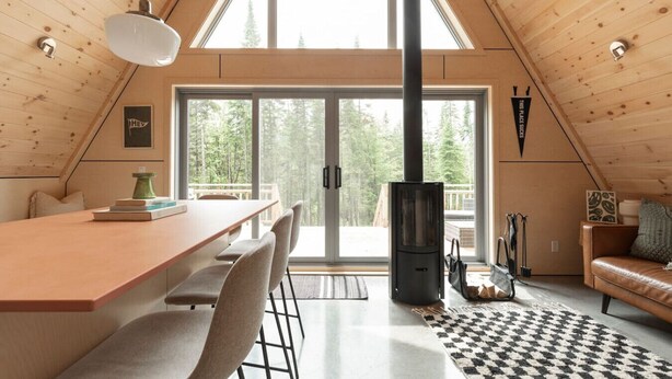 A cozy cabin interior with a dining area, a fireplace, and a large window offering a view of the forest. - Canadian Classic | Spa • Sauna • Fire • Forest (Sainte-Lucie-des-Laurentides)