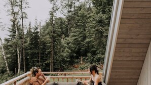 A wooden deck with wicker furniture, a railing, and a forest backdrop. - Canadian Classic | Spa • Sauna • Fire • Forest (Sainte-Lucie-des-Laurentides)