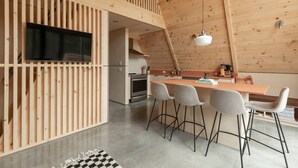 A modern kitchen with a wooden ceiling, a dining area with bar stools, and a television mounted on a wooden wall. - Canadian Classic | Spa • Sauna • Fire • Forest (Sainte-Lucie-des-Laurentides)