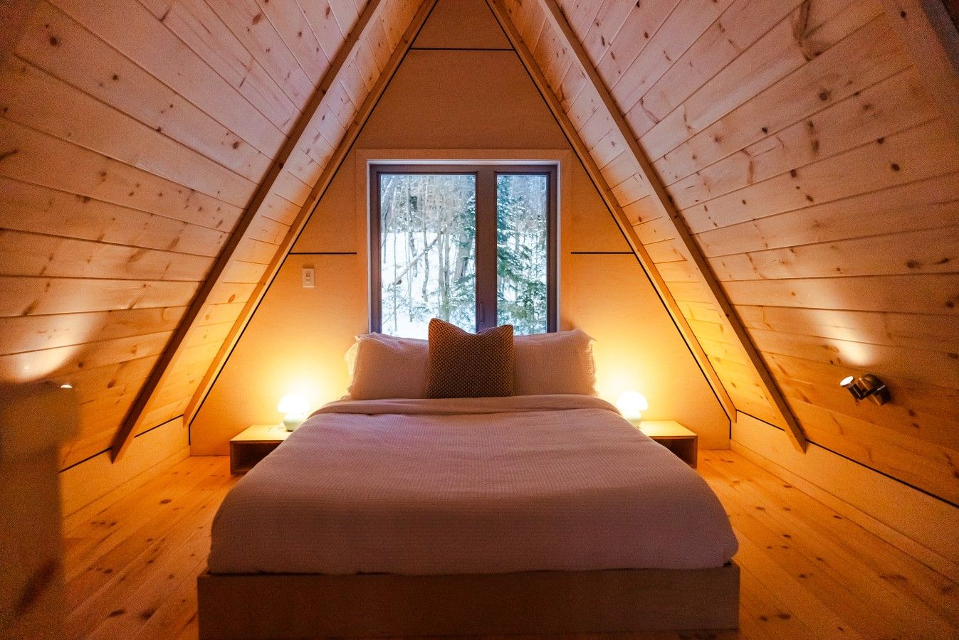 Une chambre confortable située sous les toits, avec un plafond en pente, une grande fenêtre, un lit avec des oreillers et des lampes de chevet.