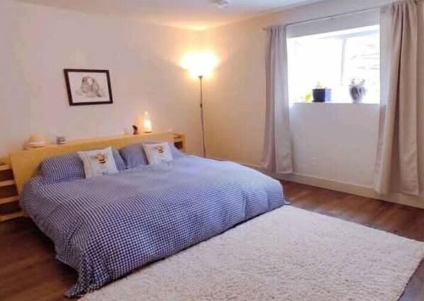 Un dormitorio con cama, mesita de noche, lámpara, ventana con cortinas e imagen enmarcada en la pared.