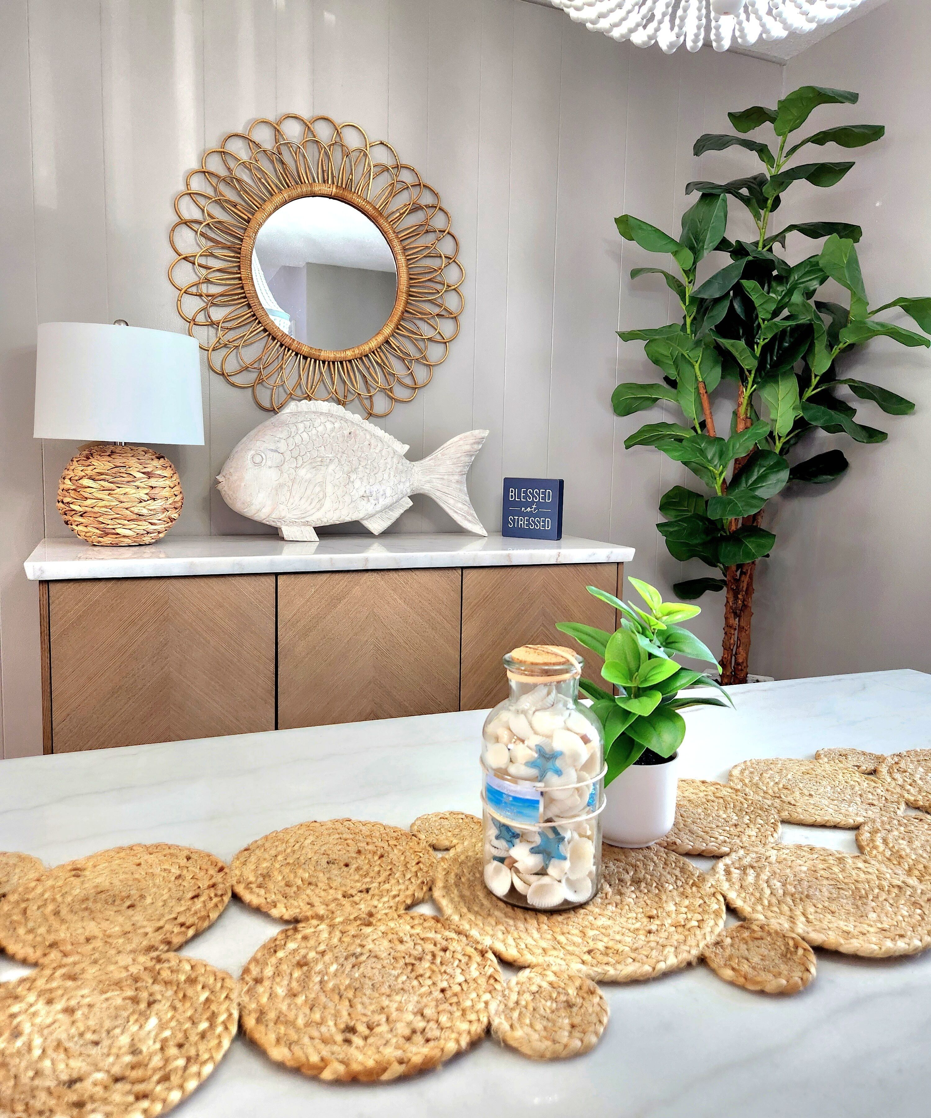 Una habitación con una mesa de mármol blanco, una escultura decorativa de pez, una lámpara tejida, una planta en maceta y un tarro de vidrio con conchas marinas.