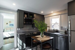 A modern kitchen with dark cabinetry, a stainless steel refrigerator, a built-in oven, and a dining area with a table and chairs.